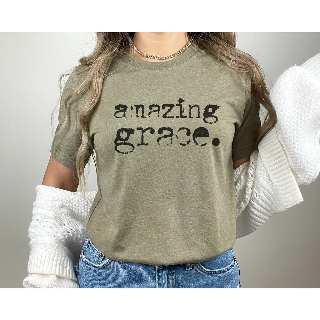 “Sage green graphic tee with distressed black typewriter text reading Amazing Grace, flat lay styled with a white hat and beaded garland.”