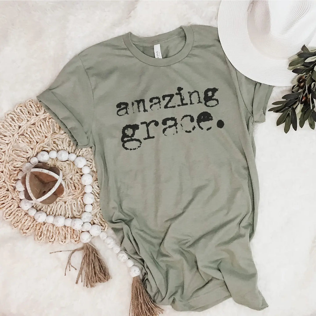 “Sage green graphic tee with distressed black typewriter text reading Amazing Grace, flat lay styled with a white hat and beaded garland.”