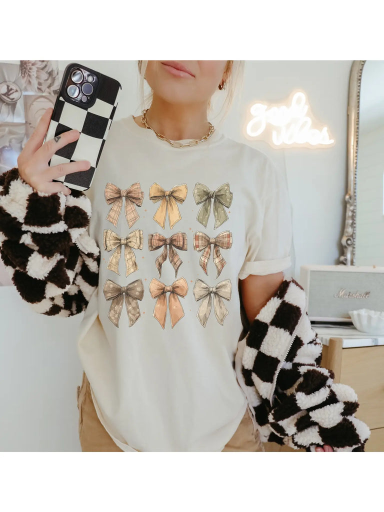 “Cream-colored graphic tee with rows of vintage plaid and neutral bows, modeled with a checkered jacket and gold necklace.”